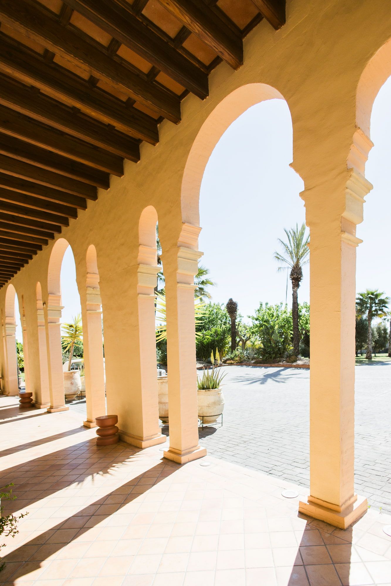 Sunlit Spanish colonial arcade with white arched columns, wooden ceiling, and palm trees visible through the archways.