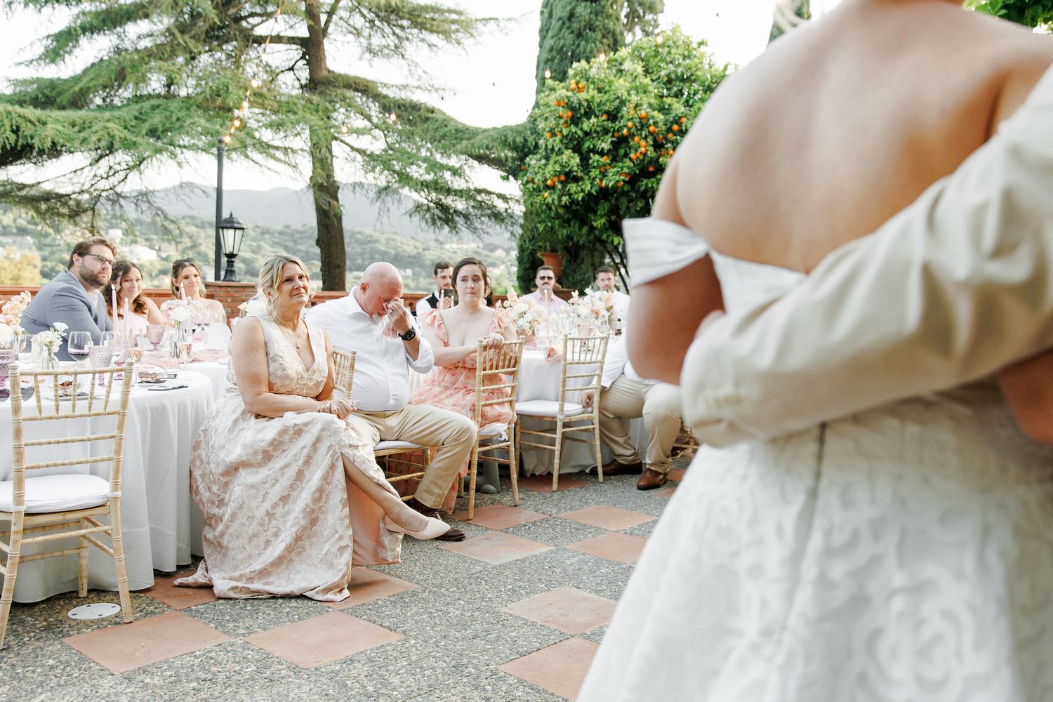 Bride in elegant white off-shoulder gown stands before seated wedding guests at an outdoor garden reception.