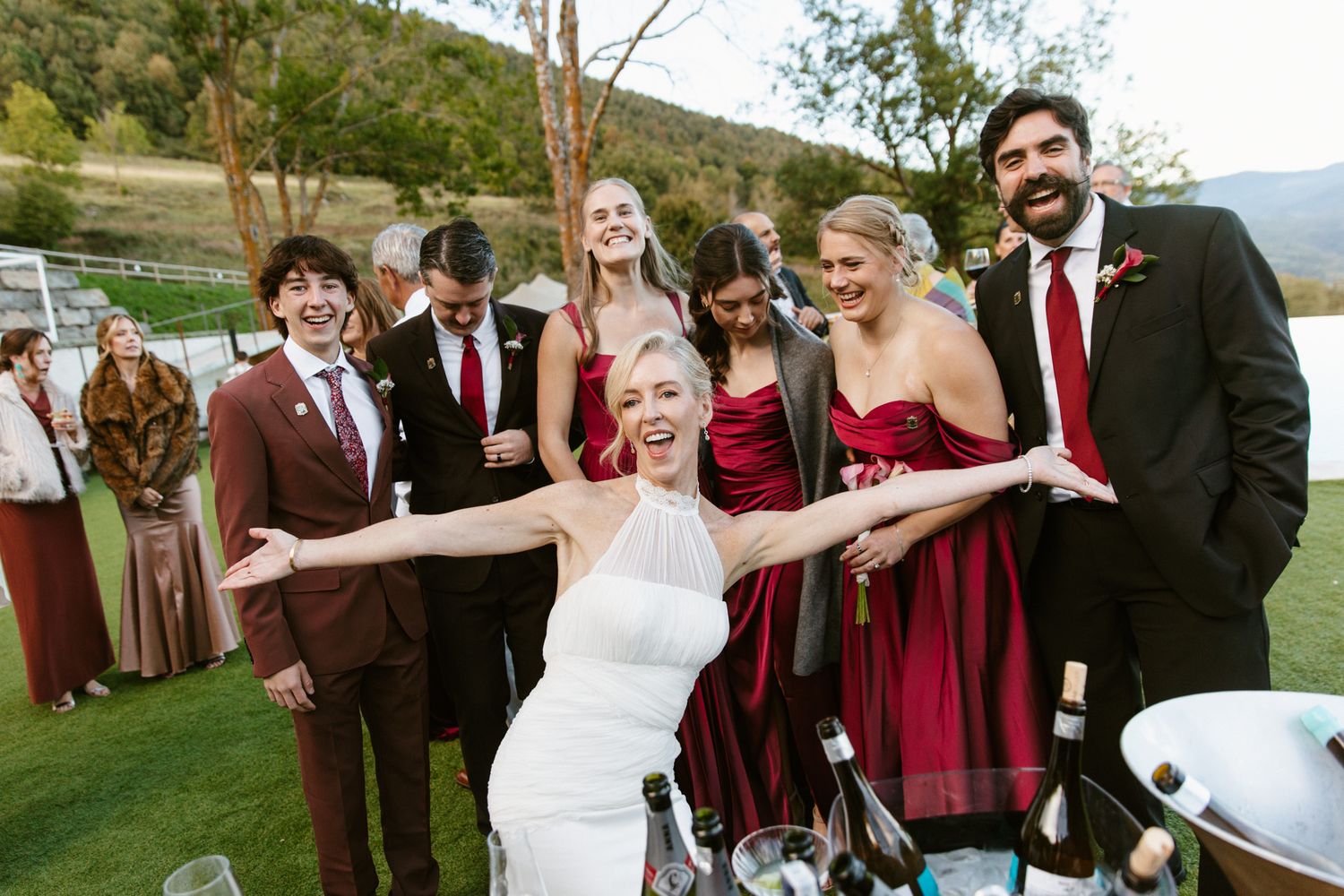 Smiling bride in a white dress leans forward with arms wide beside champagne bottles as groomsmen and bridesmaids in burgundy celebrate.