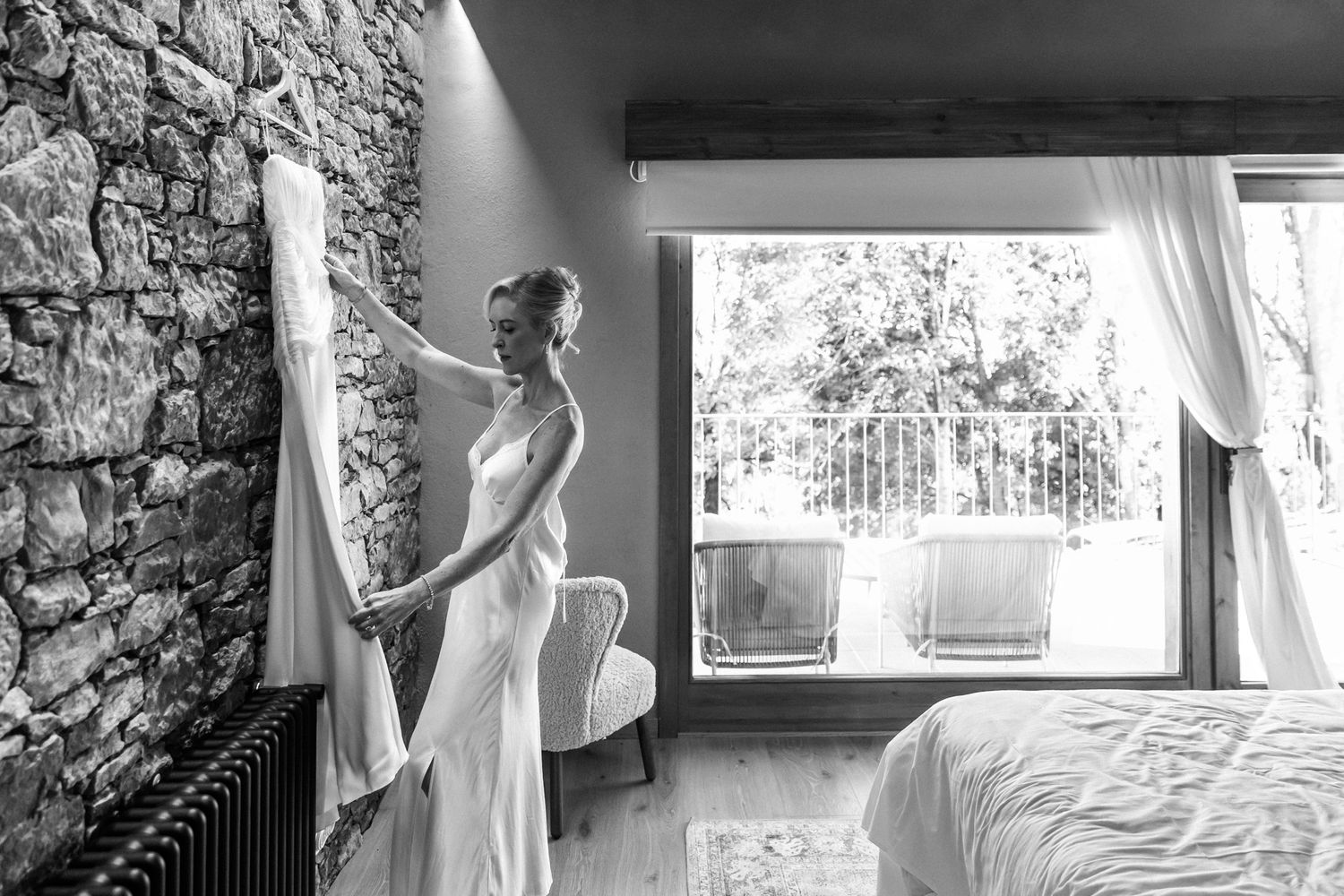 Bride in an elegant white gown adjusts her wedding dress hanging beside a rustic stone wall in a sunlit bedroom.