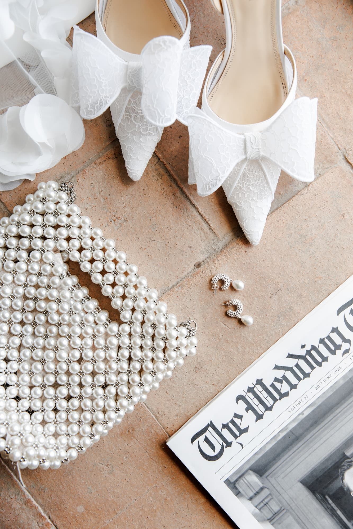 White bridal heels with bow detail, pearl beaded clutch, drop earrings, and The Wedding Times magazine on a terracotta floor.