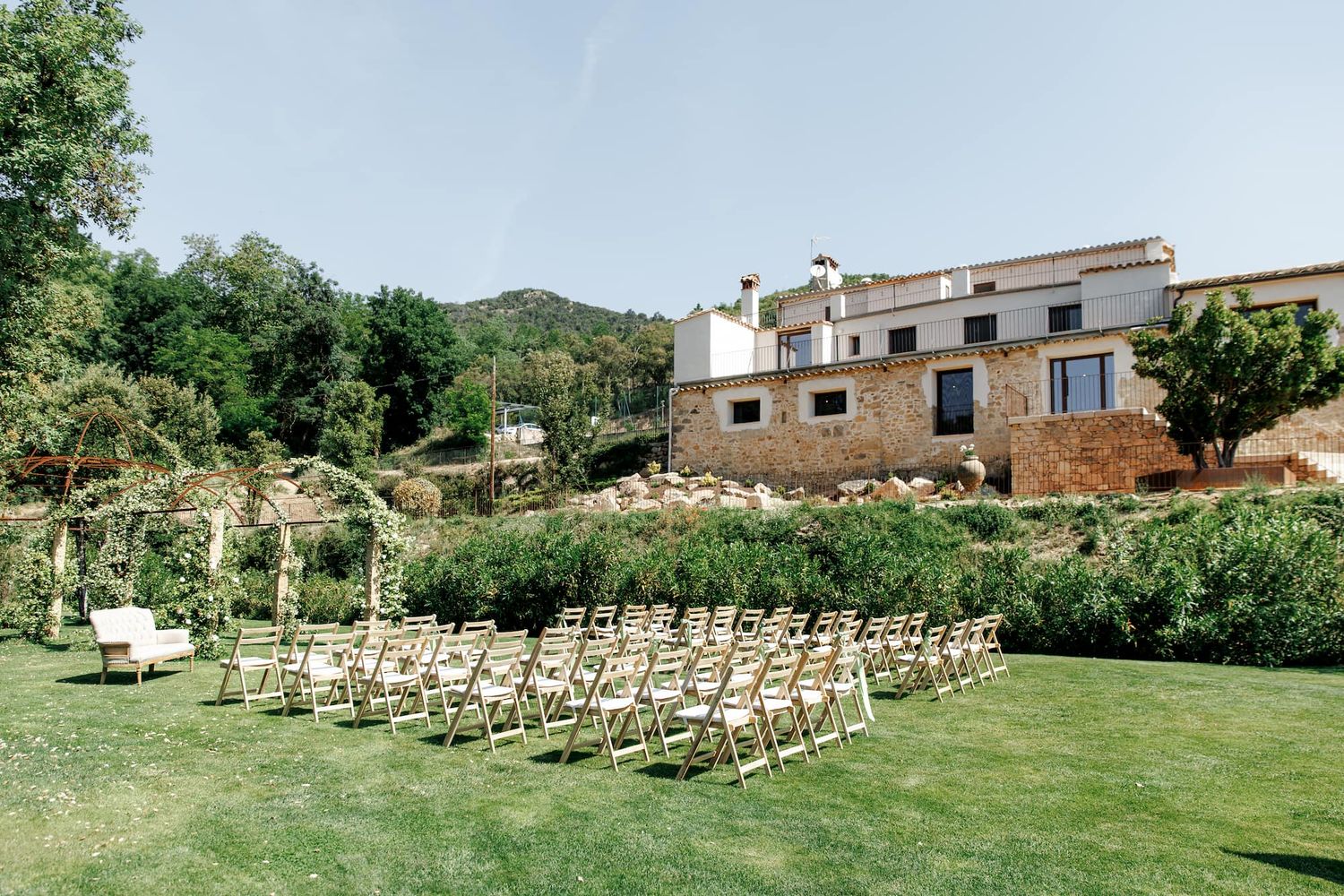 Outdoor wedding ceremony setup with wooden chairs on green lawn beside rustic stone villa surrounded by lush gardens.