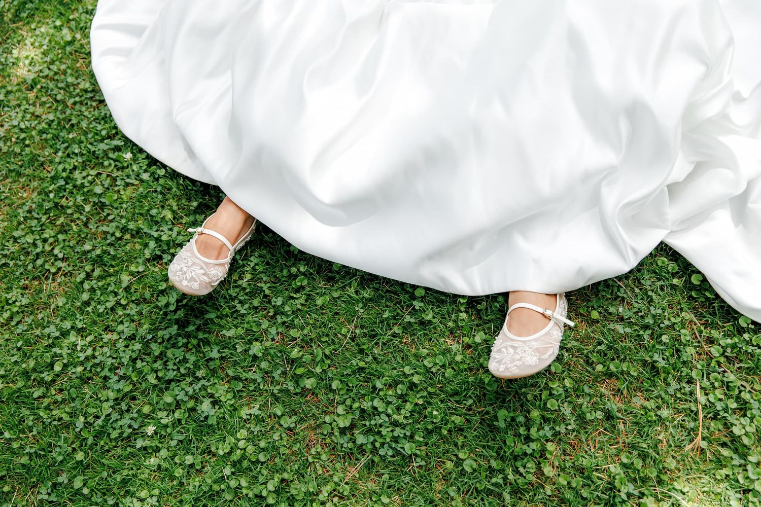 Bride wearing white lace flats peeking beneath a flowing white wedding gown on lush green grass.