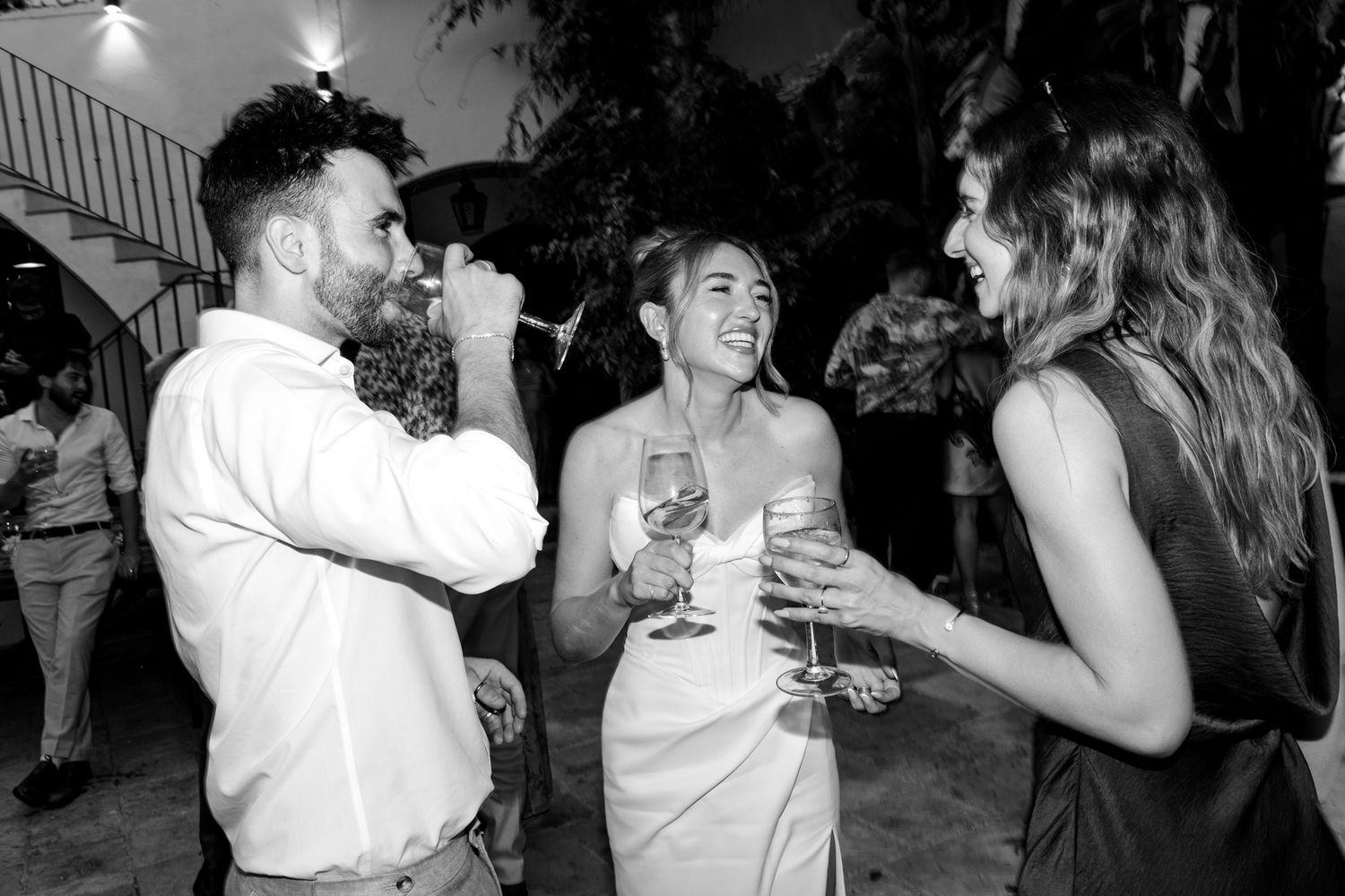 Black and white photo of three people laughing and drinking wine at an outdoor wedding party at Masia Victoria