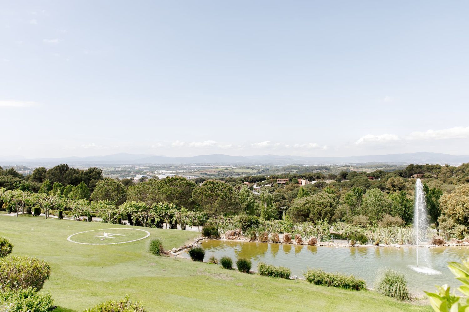 Panoramic view of lush green gardens with a fountain lake, manicured lawns, and rolling hills under a clear sky.