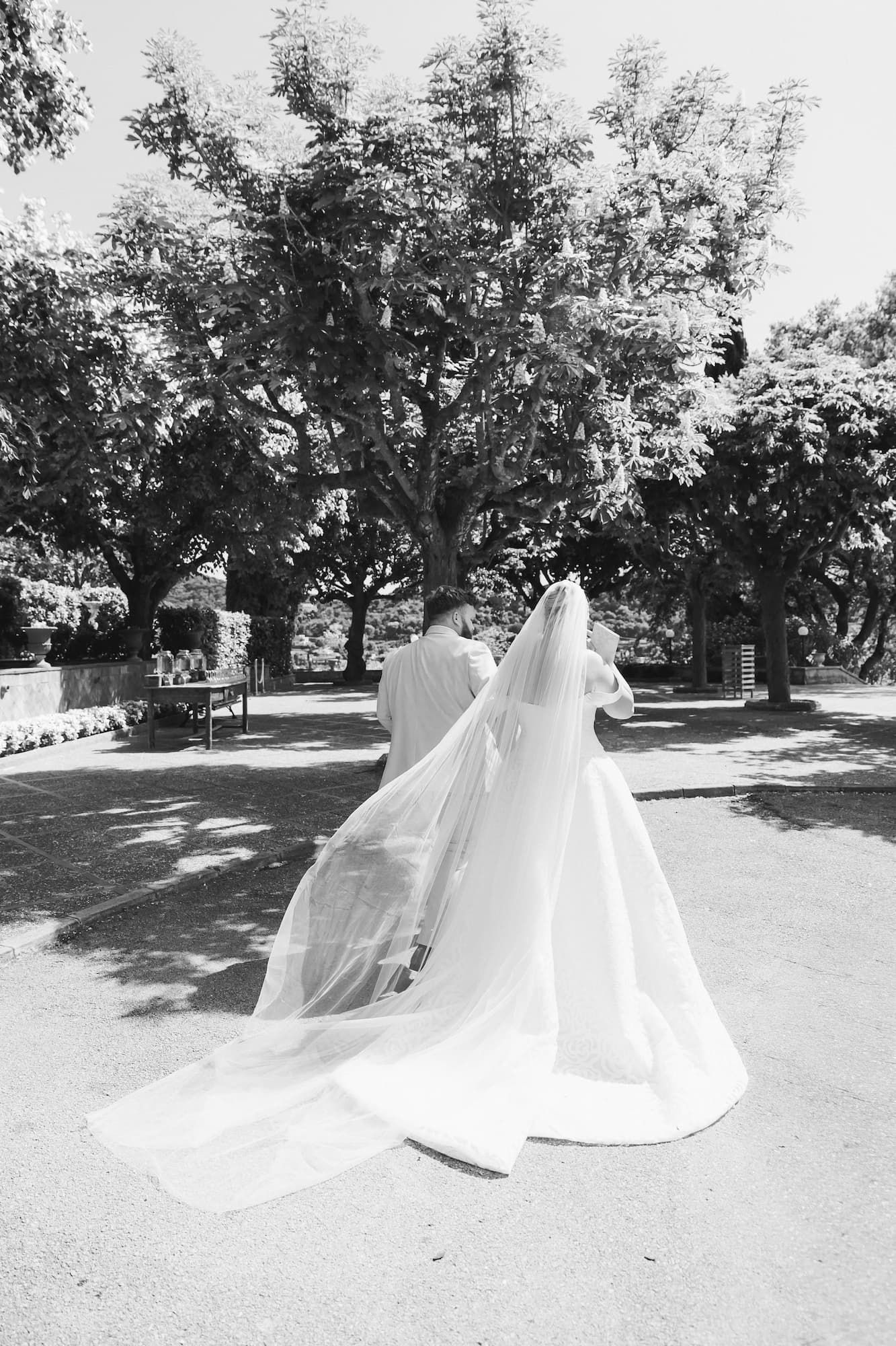 Black and white photo of a bride in a flowing gown and veil walking with groom through a sunlit park.