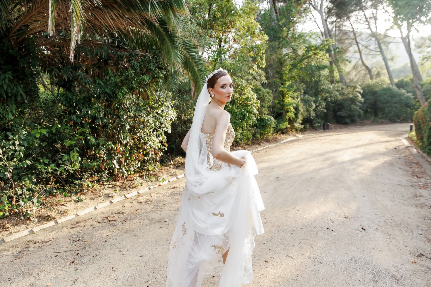A graceful figure in a flowing white wedding dress walks along a sunlit dirt path surrounded by lush greenery.