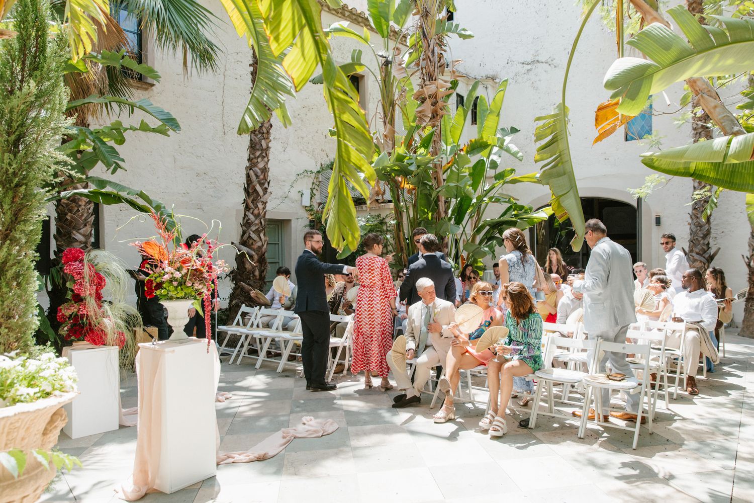 Outdoor wedding ceremony in a tropical courtyard with white chairs, palm trees, and guests gathered under bright sunlight.