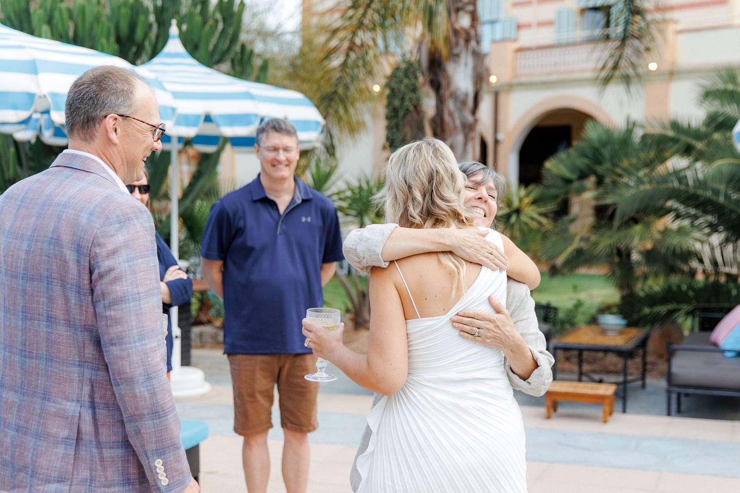 Bride in white dress hugging a guest outdoors at an elegant venue with blue striped umbrella and palm trees.
