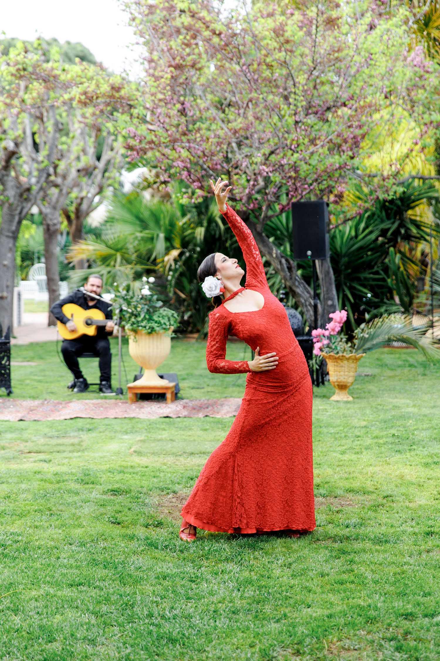 Flamenco dancer in red dress performs outdoors with guitarist accompaniment in lush garden setting.