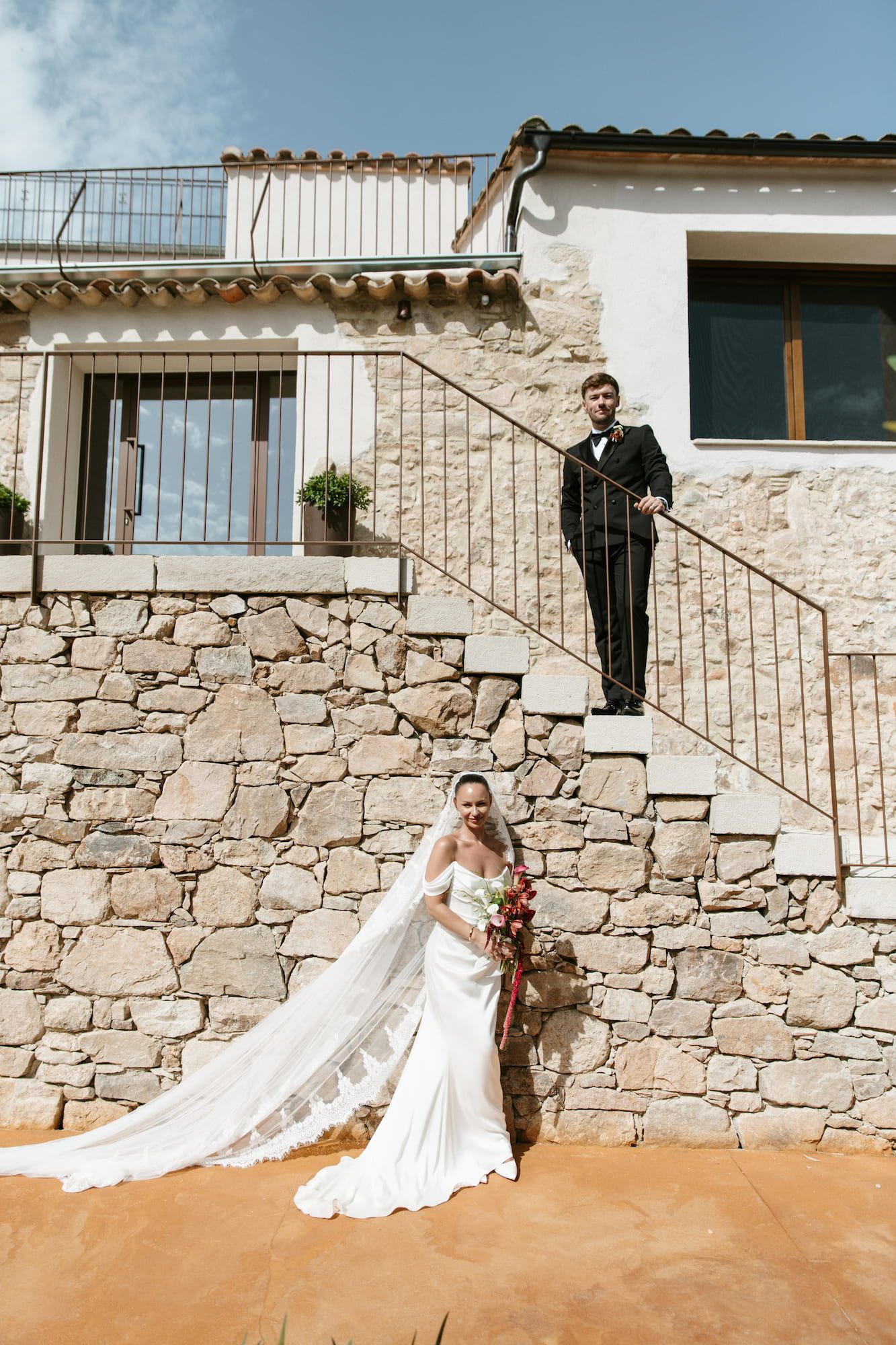Bride in white gown with veil stands by stone wall as groom in black tuxedo poses on staircase.