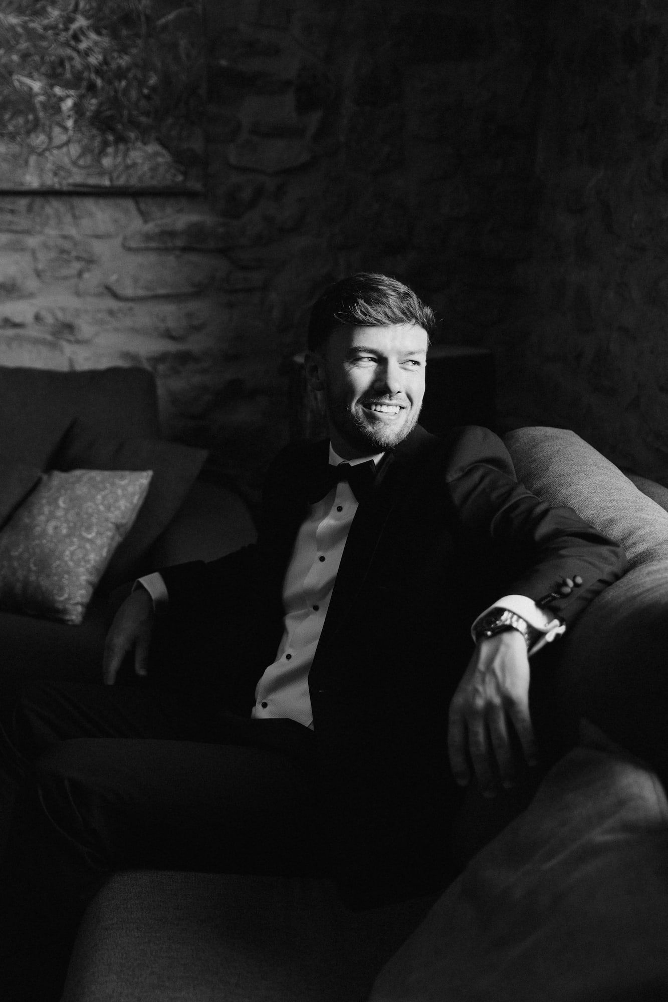 Black and white photo of a smiling groom in a tuxedo with bow tie, relaxing on a sofa in a rustic stone venue.