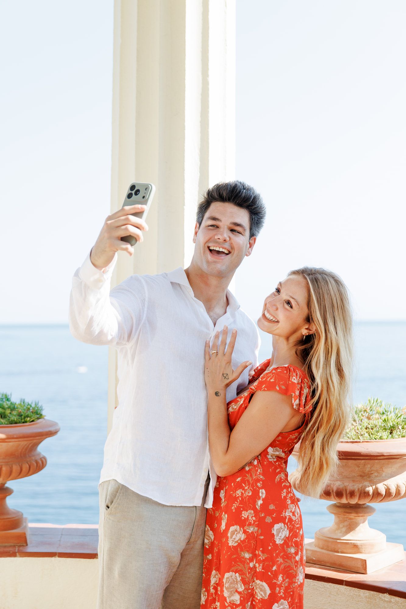 A joyful couple takes a selfie on a sunny terrace overlooking the sea, as the woman shows off her engagement ring.