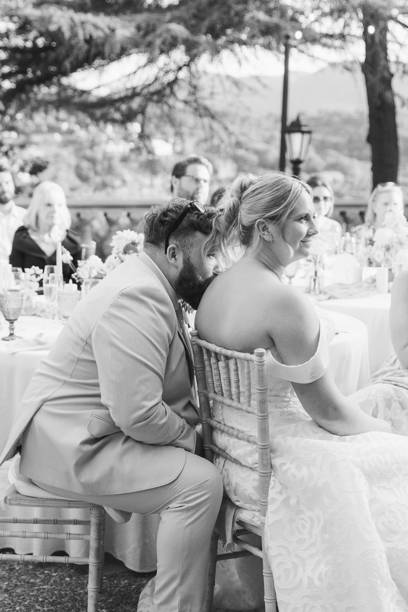 Black and white photo of bride and groom seated at outdoor wedding reception table, groom leaning toward bride.