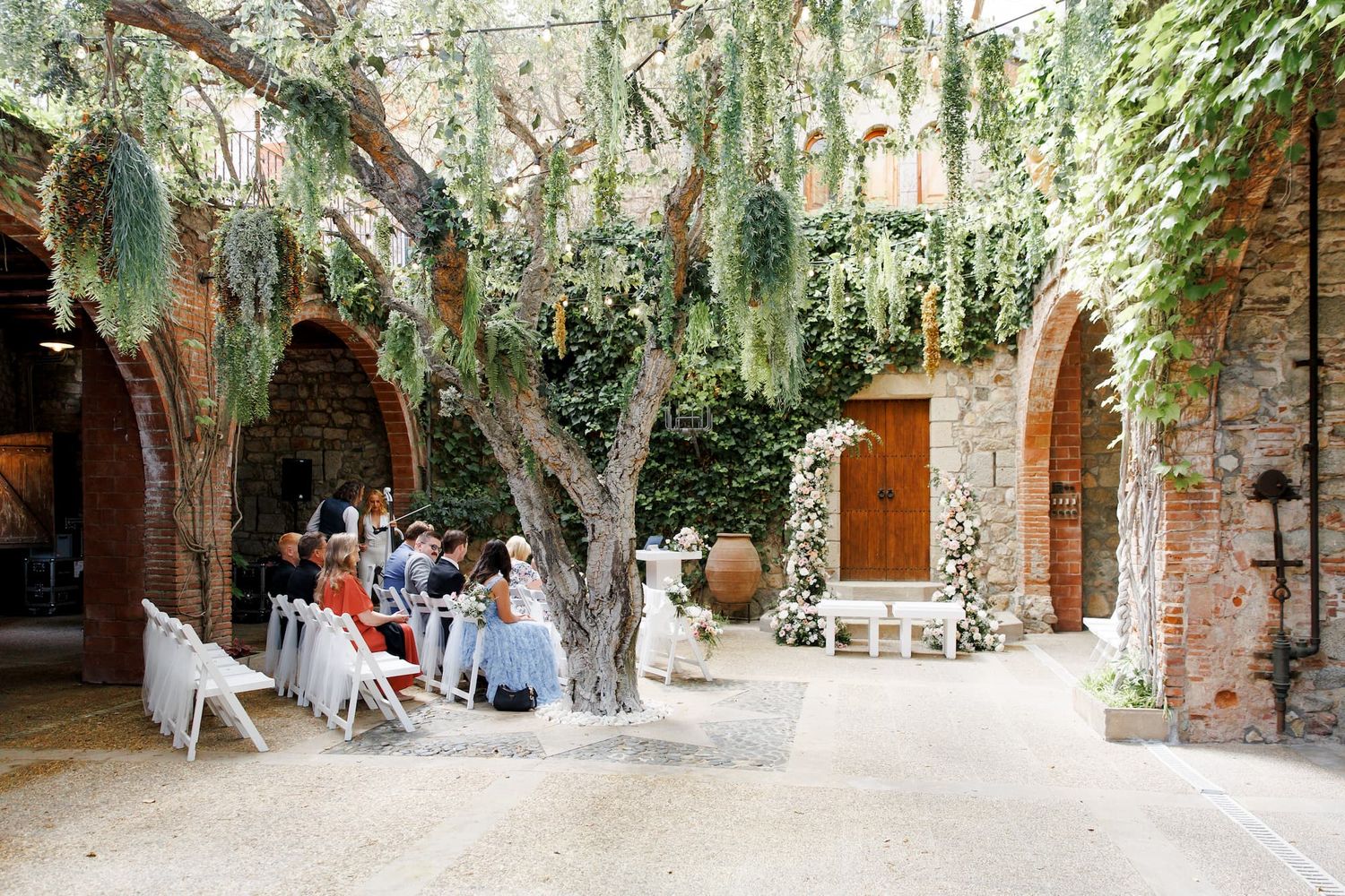 Romantic outdoor wedding ceremony in a rustic stone courtyard with arched walls, lush greenery, and a majestic tree centerpiece.