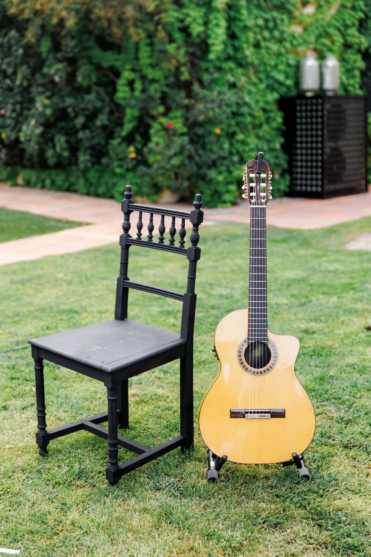 Black wooden chair and acoustic classical guitar on guitar stand in lush green garden setting.
