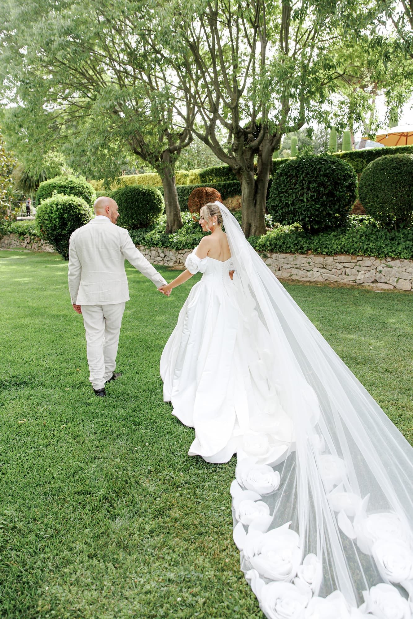 Bride in a long white gown and veil holds hands with groom in beige suit walking through a lush green garden.