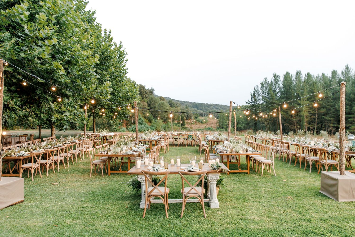 Outdoor wedding reception in Barcelona on green lawn with string lights, wooden chairs, and long rustic tables surrounded by tall trees.