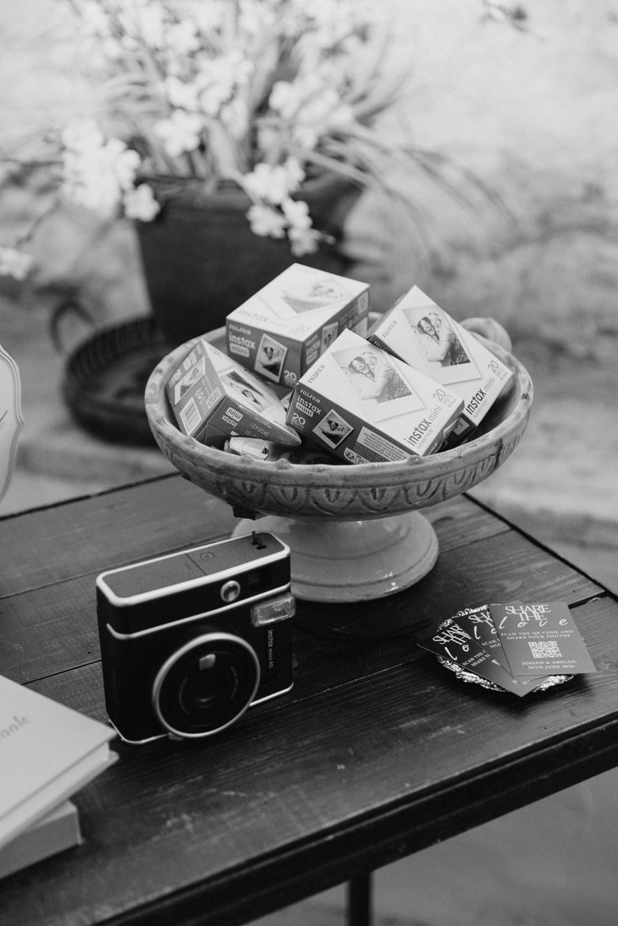 Black and white photo of an instant camera beside a bowl of film boxes on a wooden table with flowers in background.