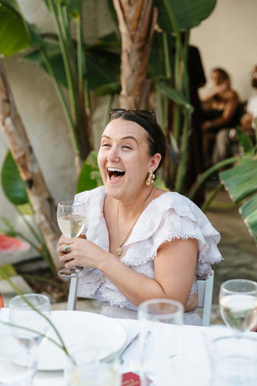 A joyful woman in a white dress laughs while holding a wine glass at an outdoor garden dining event.