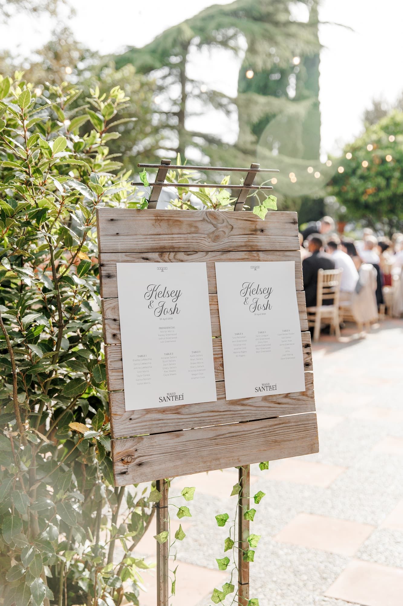 Rustic wooden easel displaying wedding seating charts for Kelsey and Josh at an outdoor garden reception.