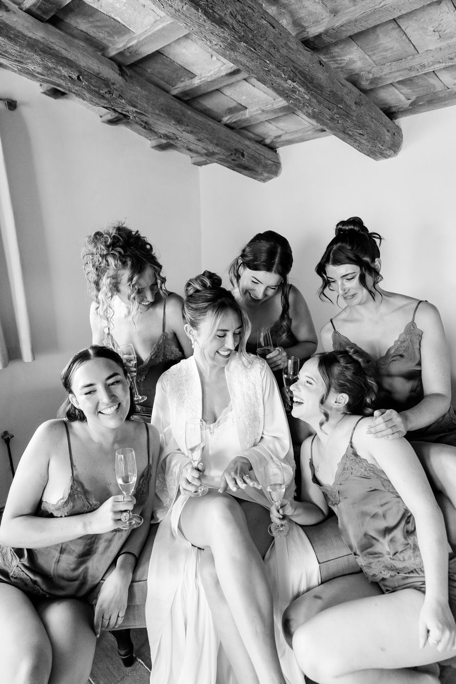 Bride in white robe laughs with five bridesmaids in silk slips while celebrating with champagne.