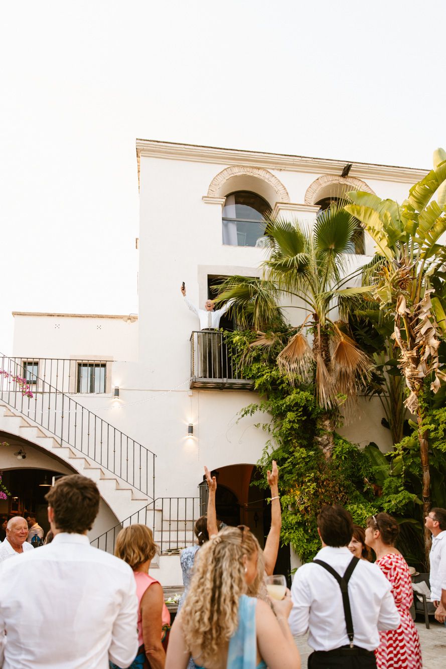 Guests gather in a sunlit Mediterranean courtyard, looking up at Masia Victoria with balcony, palm trees, and iron staircase.