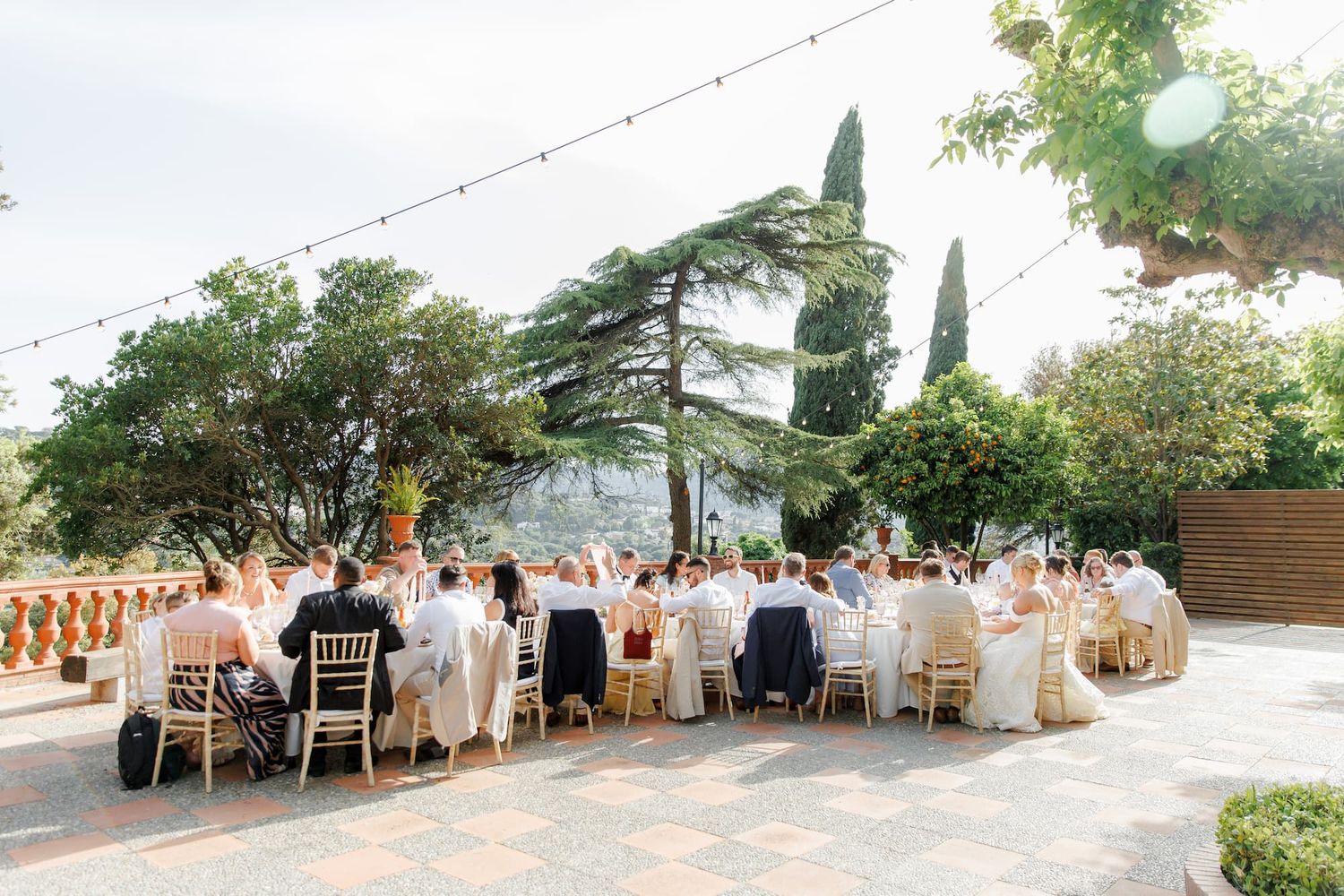 Elegant outdoor wedding reception on a sunlit terrace with string lights, lush trees, and guests seated at decorated tables.