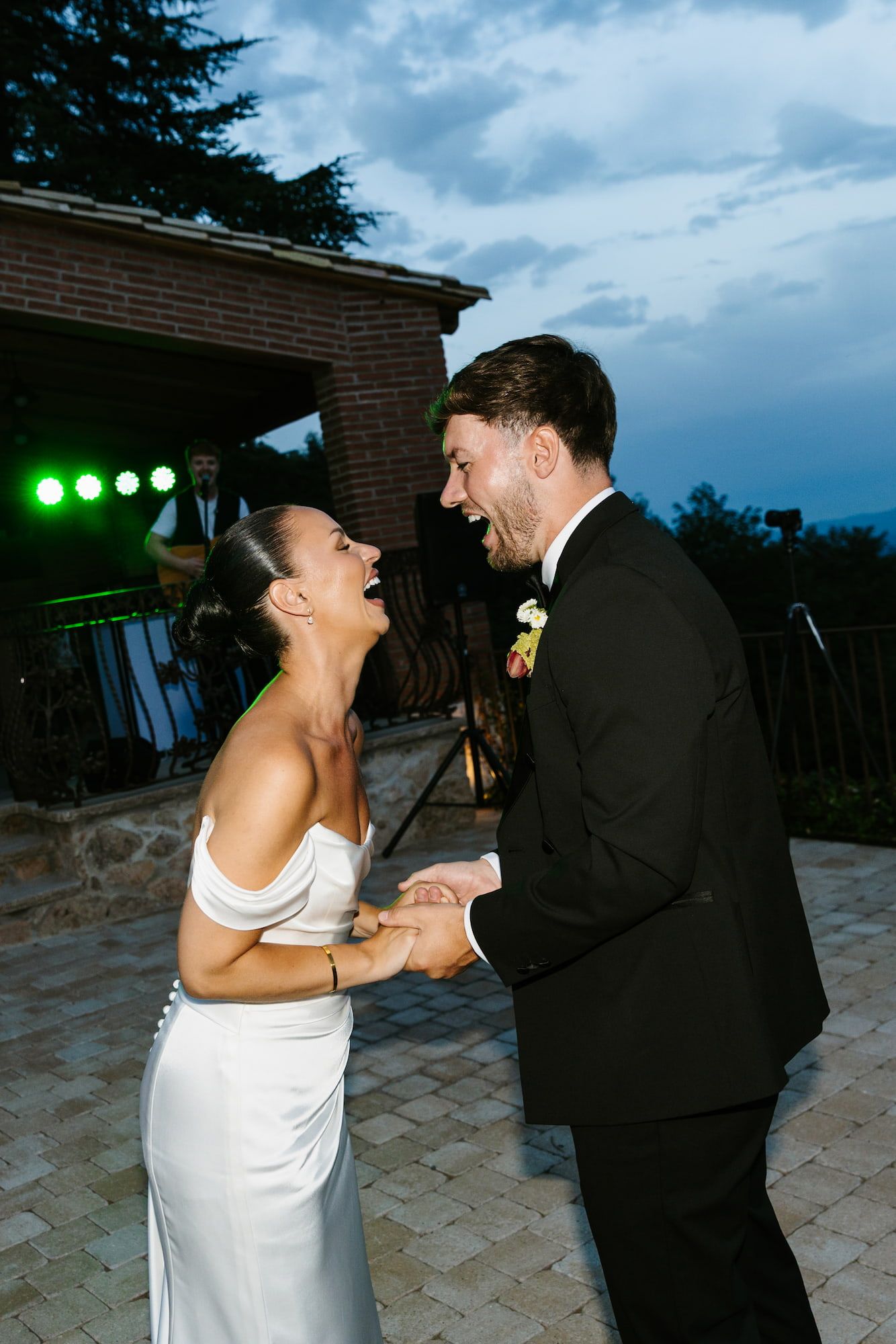 Bride and groom laughing joyfully while holding hands during their first dance at an outdoor evening wedding reception.