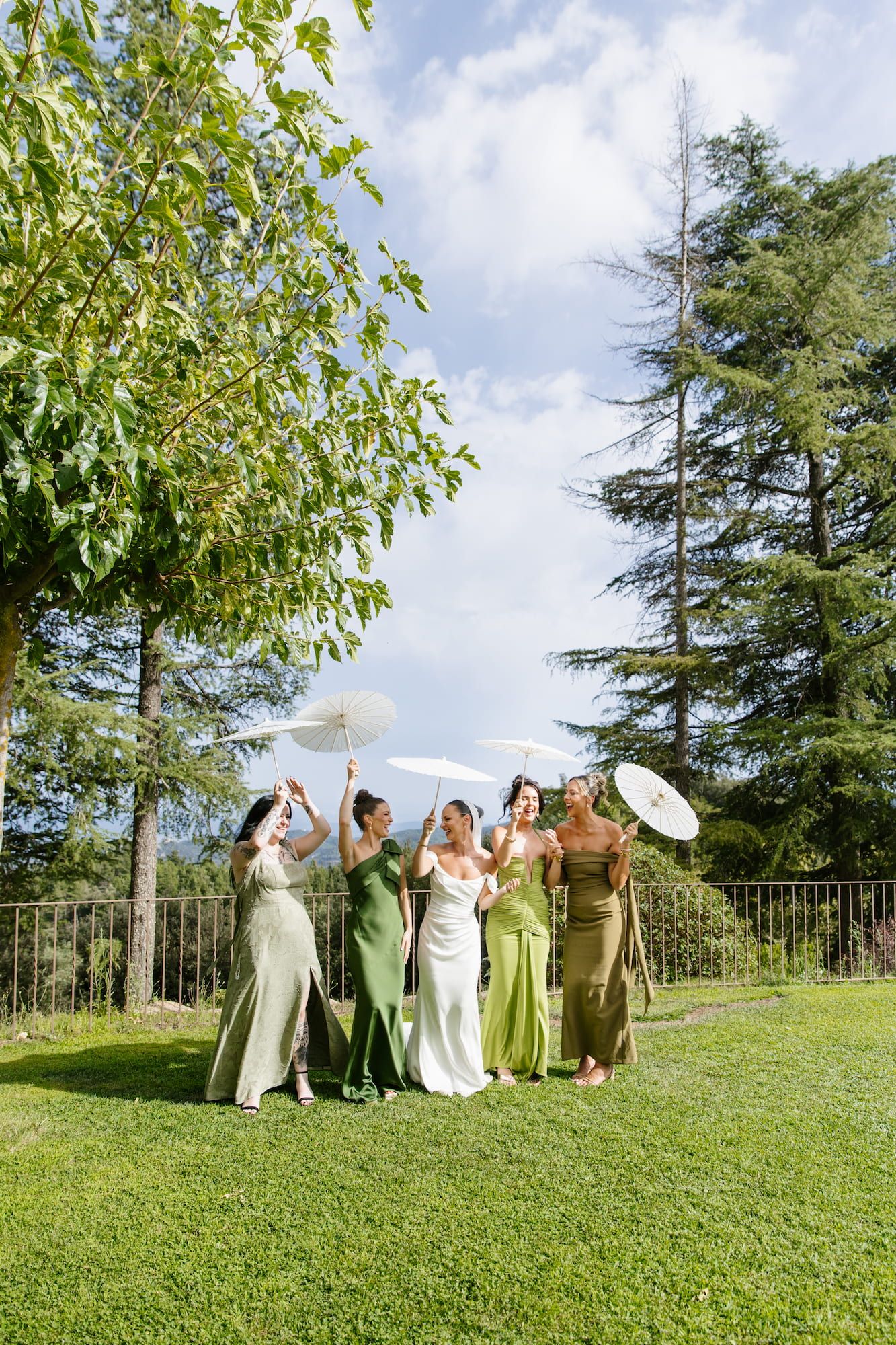 Bride and bridesmaids in green dresses holding white parasols on a sunny garden lawn.