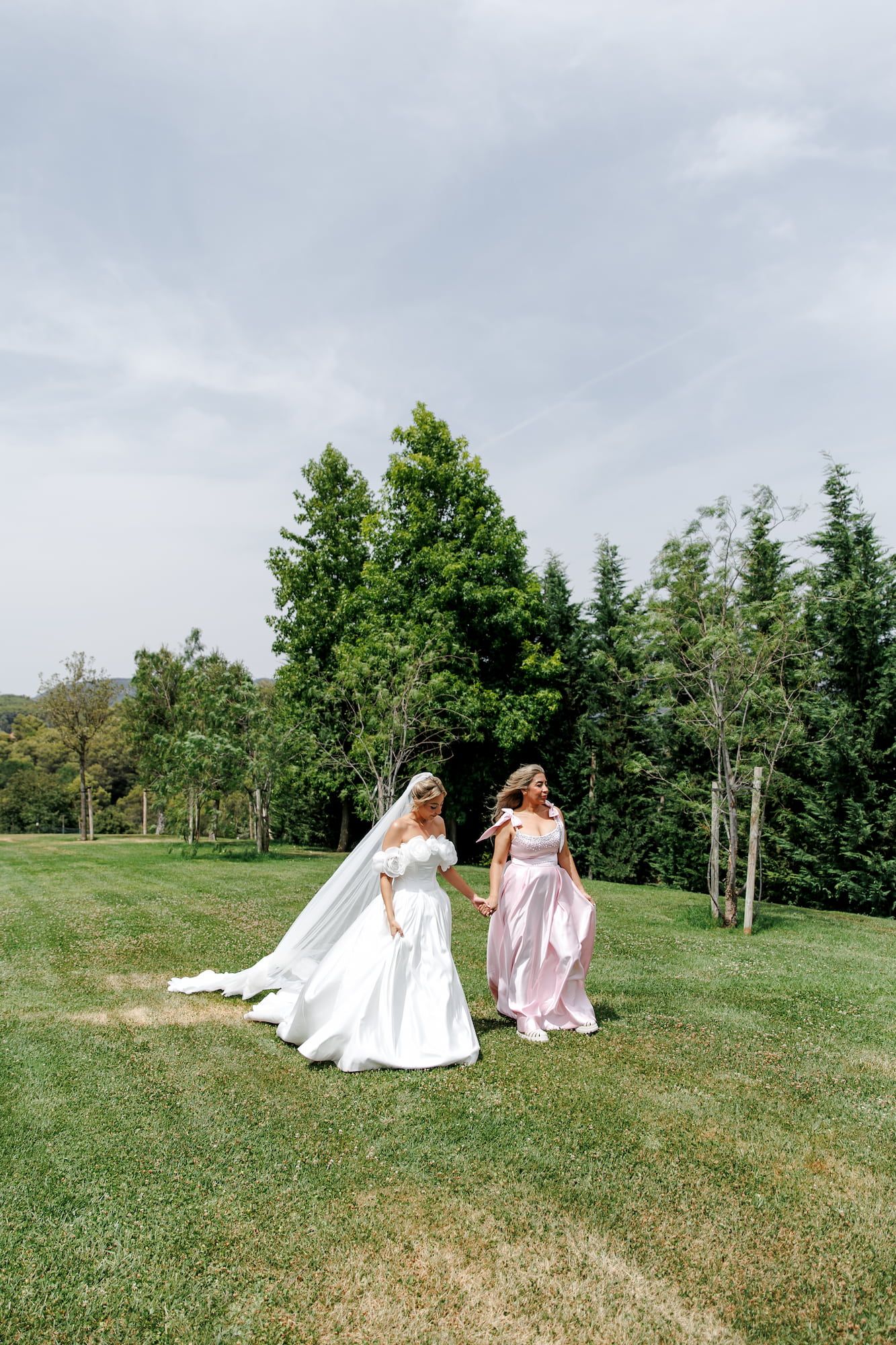 Bride in white gown and bridesmaid in pink dress walking on green lawn with trees in background.