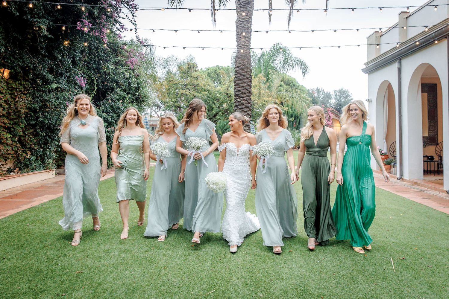 Bride in white gown walks with bridesmaids in sage green dresses outdoors at a sunny garden wedding venue in Barcelona