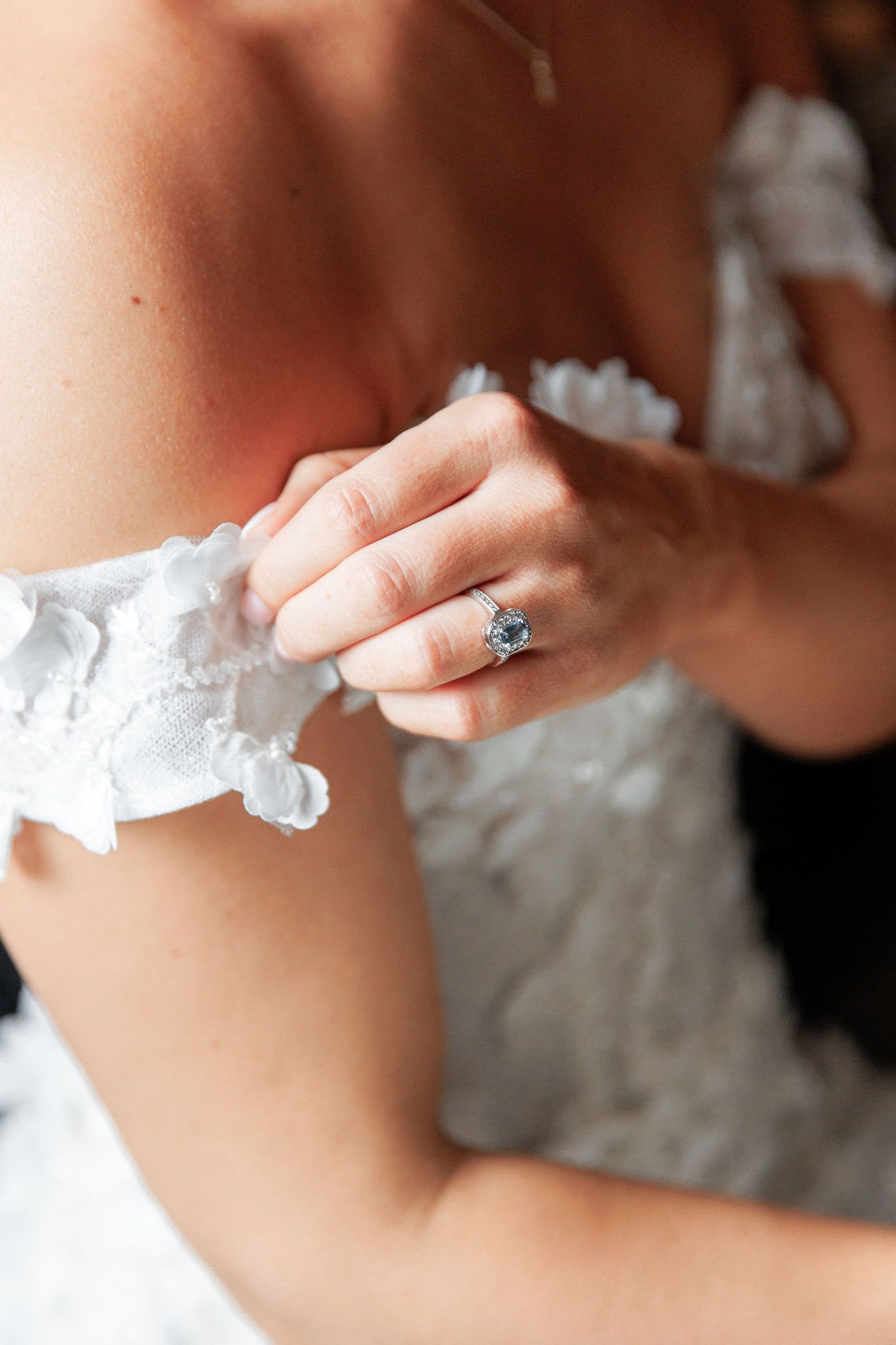 Bride wearing a white lace wedding dress adjusts her garter while showcasing a blue sapphire engagement ring.