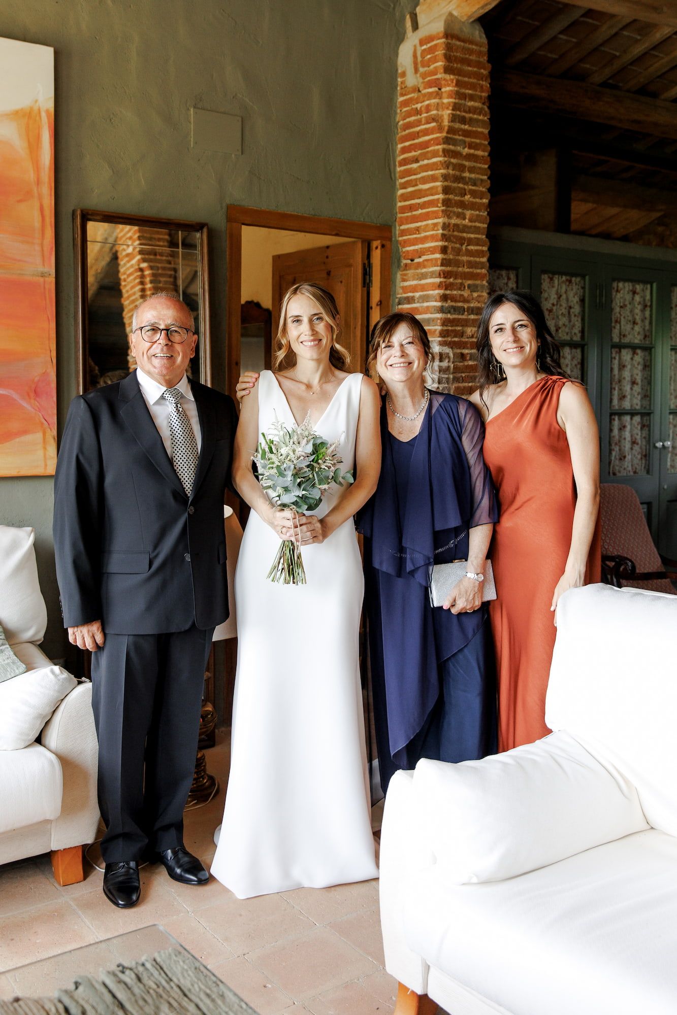 Bride in white gown holds bouquet, posing with family in navy and rust attire inside elegant venue.