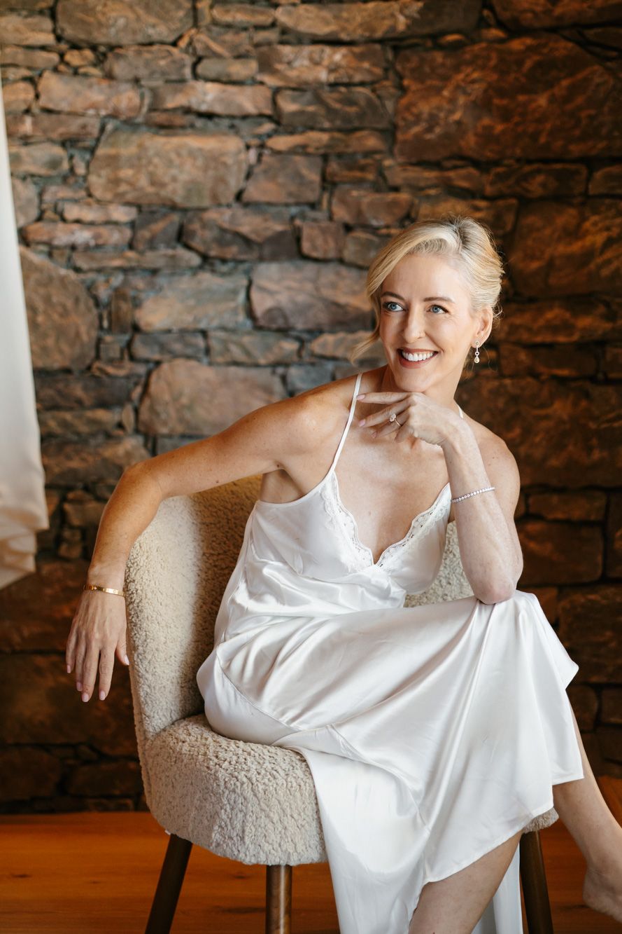 Smiling woman with short blonde hair in a white satin slip dress sits on a cream boucle chair in front of a rustic stone wall.