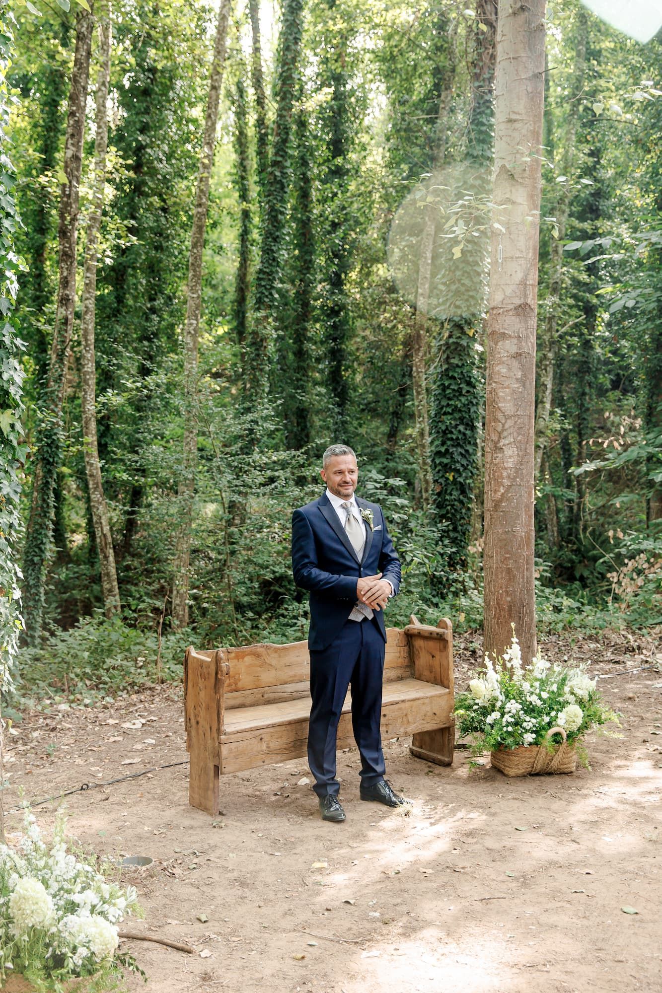 Groom in navy suit smiling at outdoor woodland wedding ceremony with rustic wooden bench and white floral arrangements.