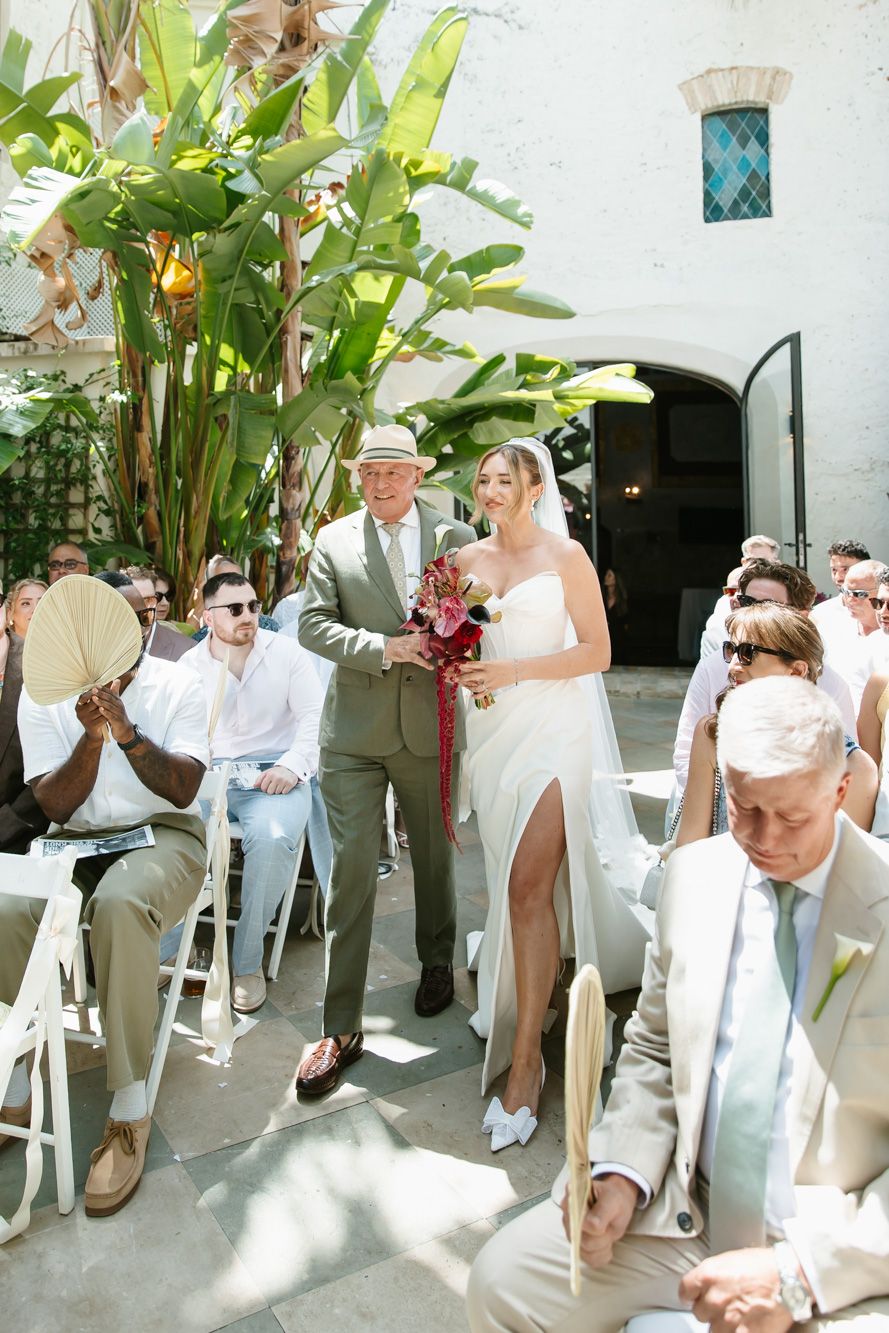 Bride walks down outdoor aisle with escort at sunny wedding ceremony, guests seated in white chairs amid tropical greenery.
