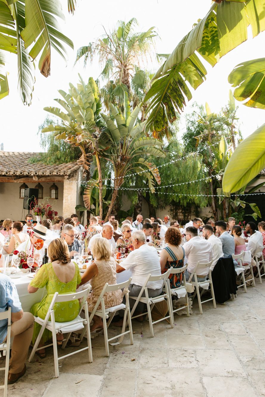 Outdoor wedding reception at Masia Victoria with guests seated at long tables under tropical palm trees with string lights.