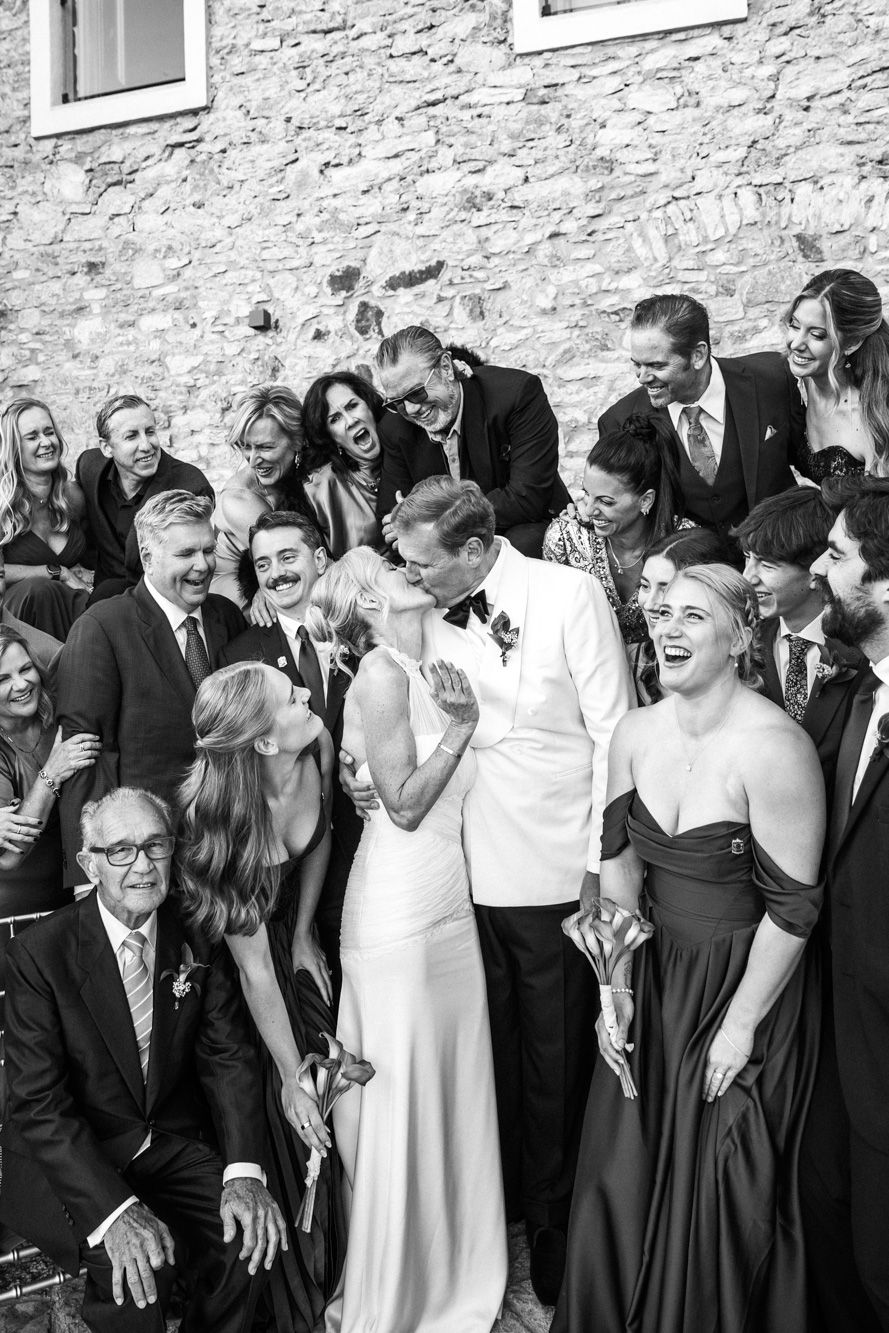 Black and white wedding photo of bride and groom kissing surrounded by laughing guests near a stone wall.