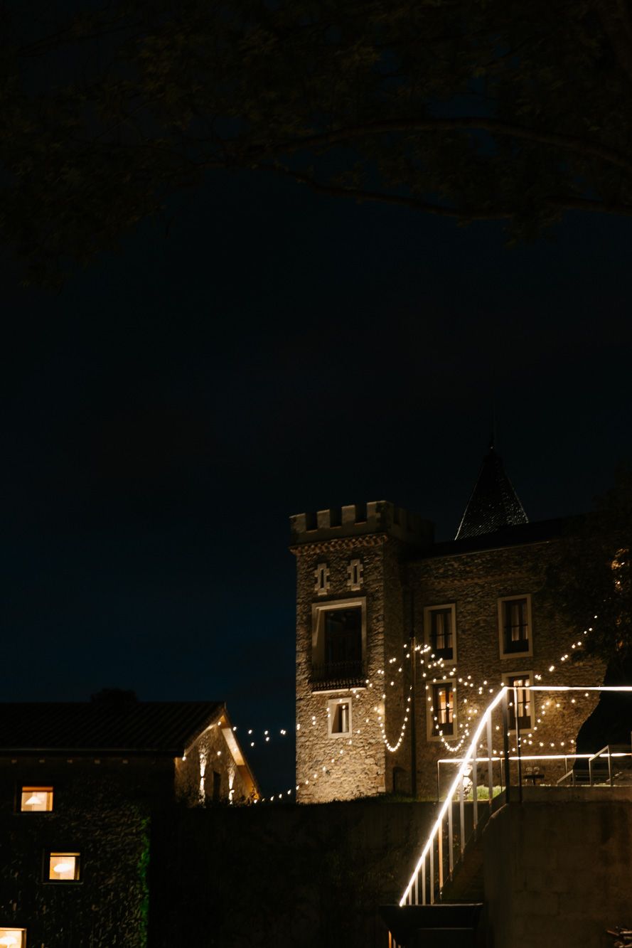 Stone castle tower with crenellations illuminated by warm string lights against a dark night sky, with glowing windows nearby.