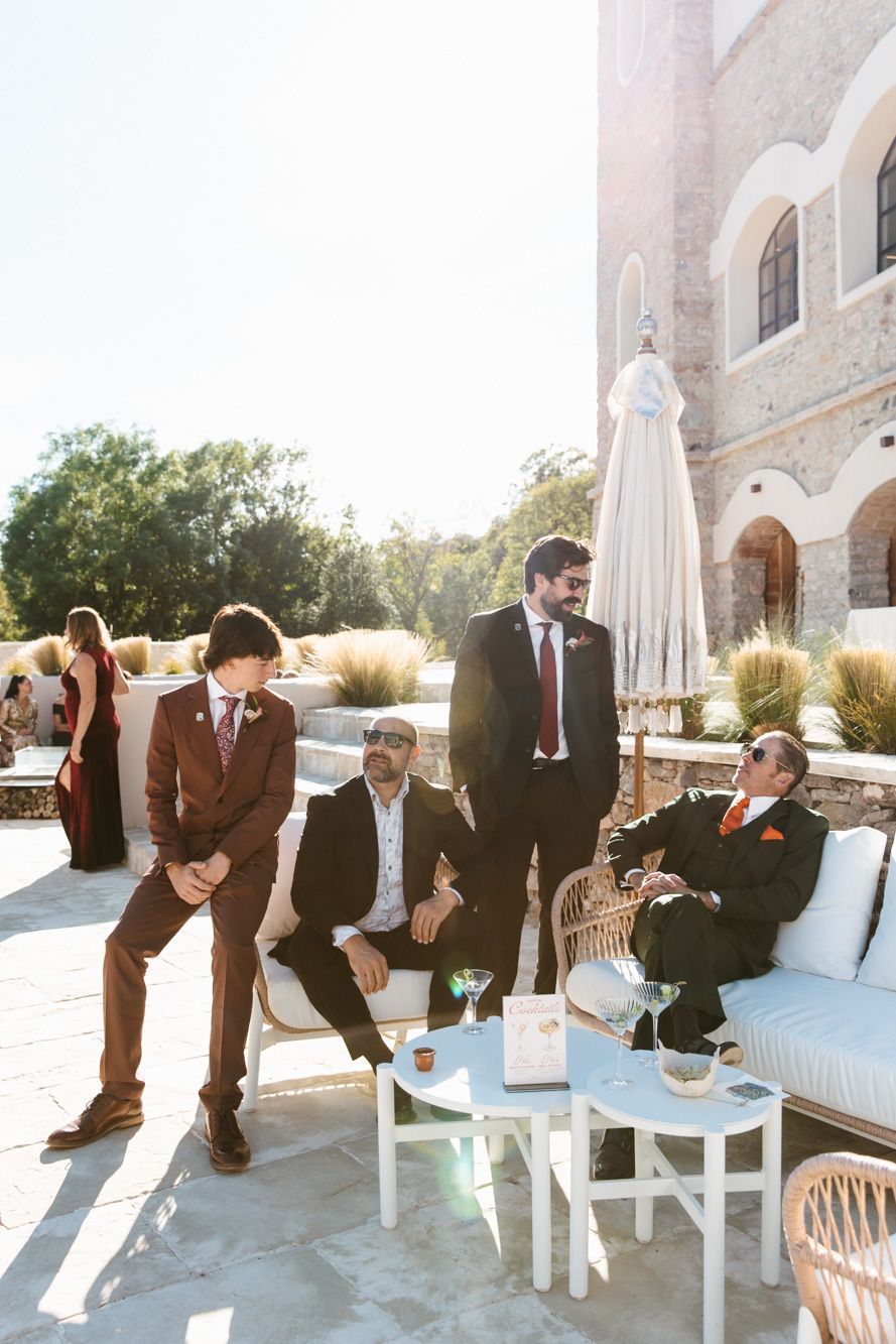 Four men in burgundy and dark suits chat and relax with cocktails on a sunny stone terrace beside a rustic building at an outdoor wedding.
