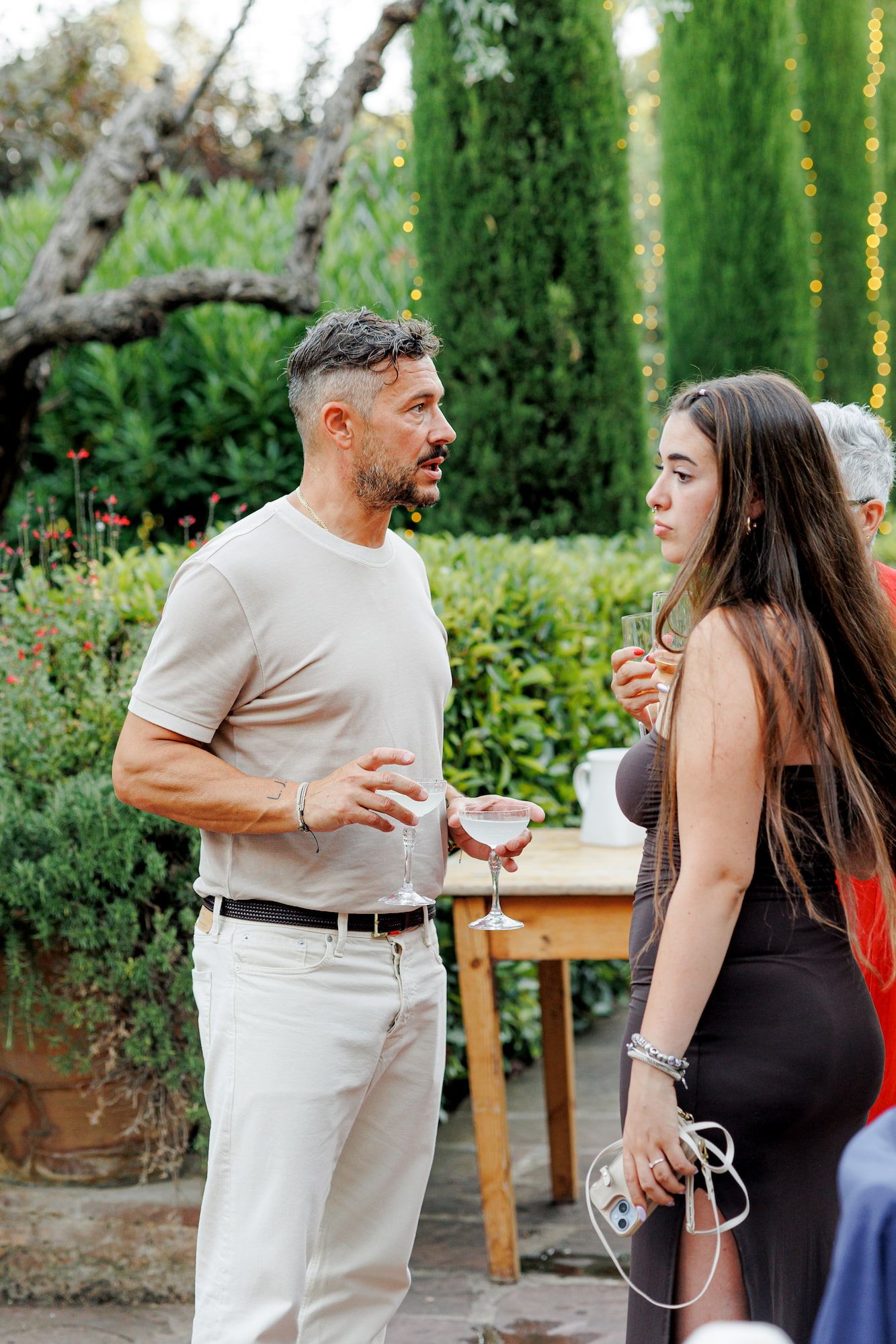 Man and woman conversing at an outdoor garden party, holding cocktails among lush greenery.