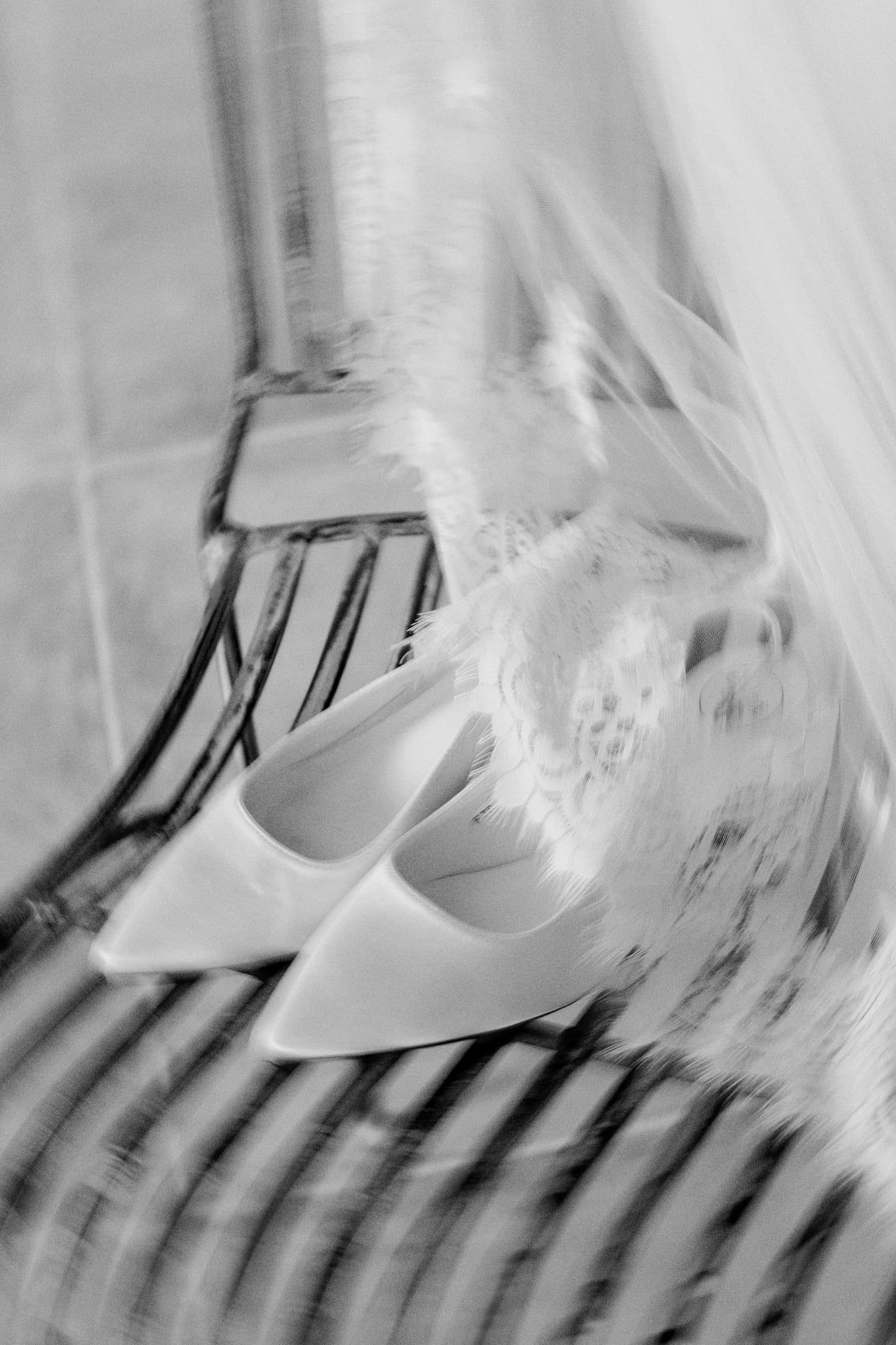 Black and white photo of white bridal heels resting on a chair beneath a flowing wedding veil.