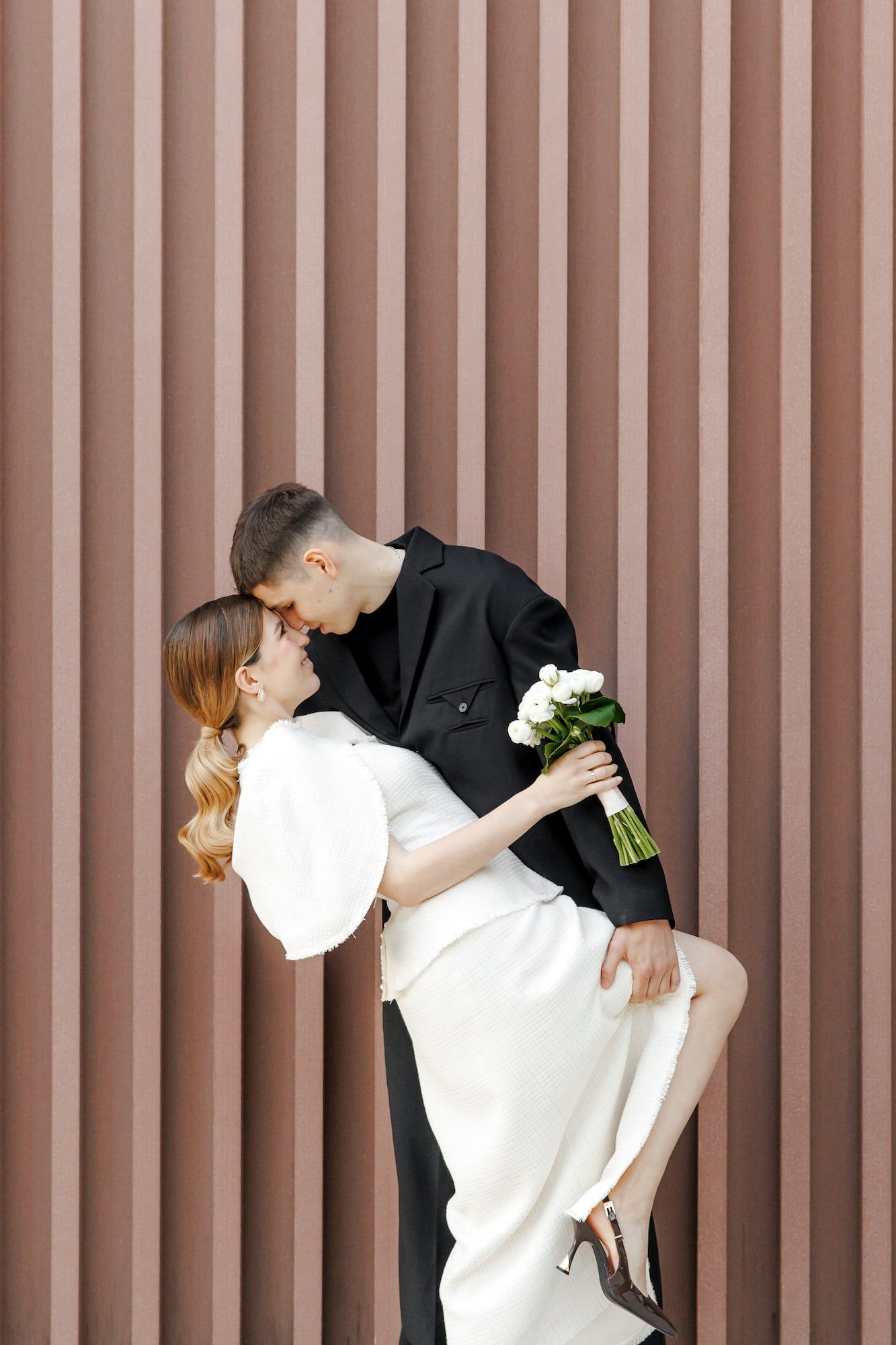 A groom in a black suit dips his bride in a white dress as they kiss, holding a white rose bouquet against a striped wall.