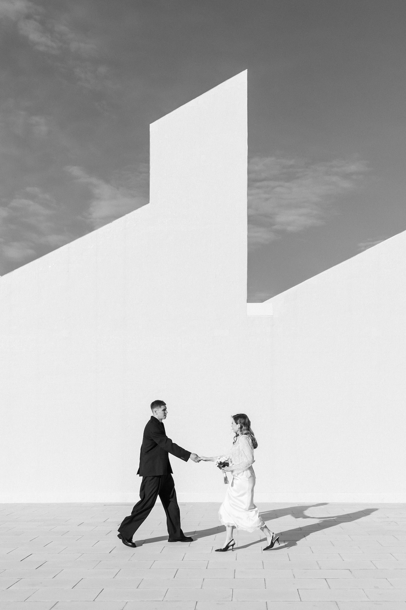 Black and white photo of a couple holding hands walking past a minimalist white architectural wall outdoors.