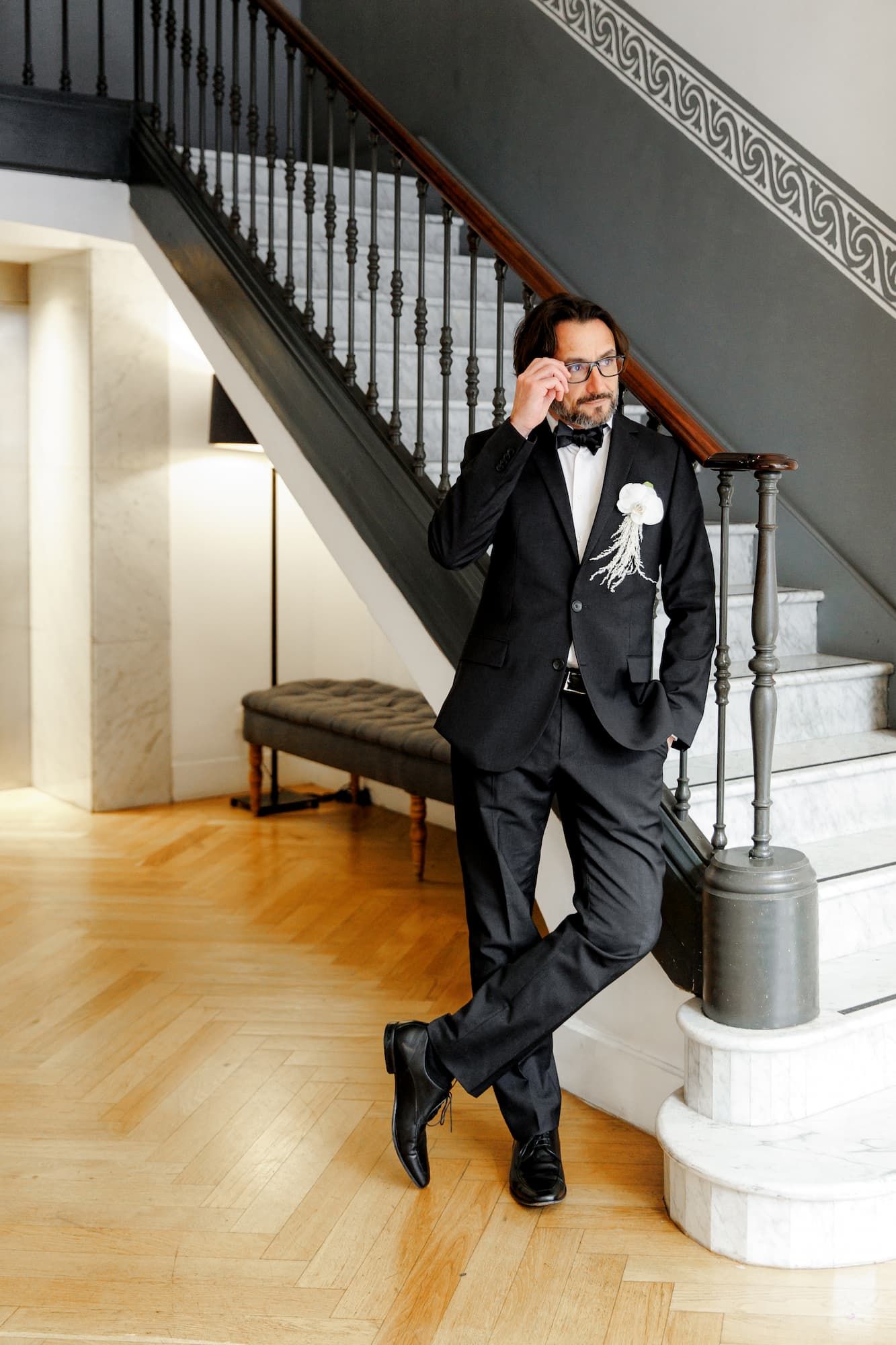 An elegant groom in a black tuxedo and bow tie poses near a curved staircase with an ornate railing.