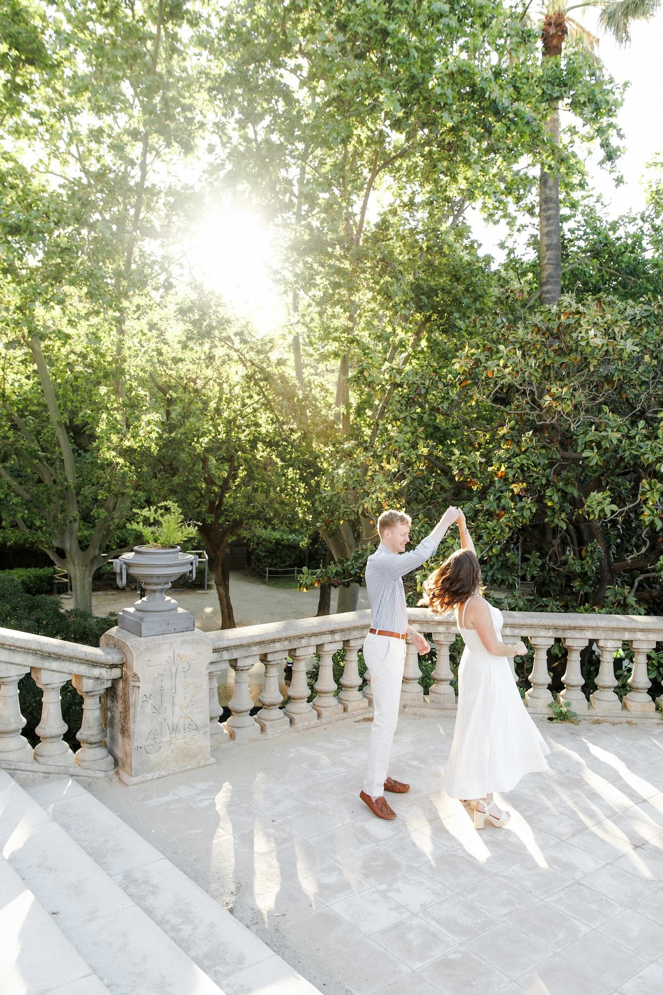 Couple dancing on sunlit stone terrace surrounded by lush green trees in an elegant garden setting.