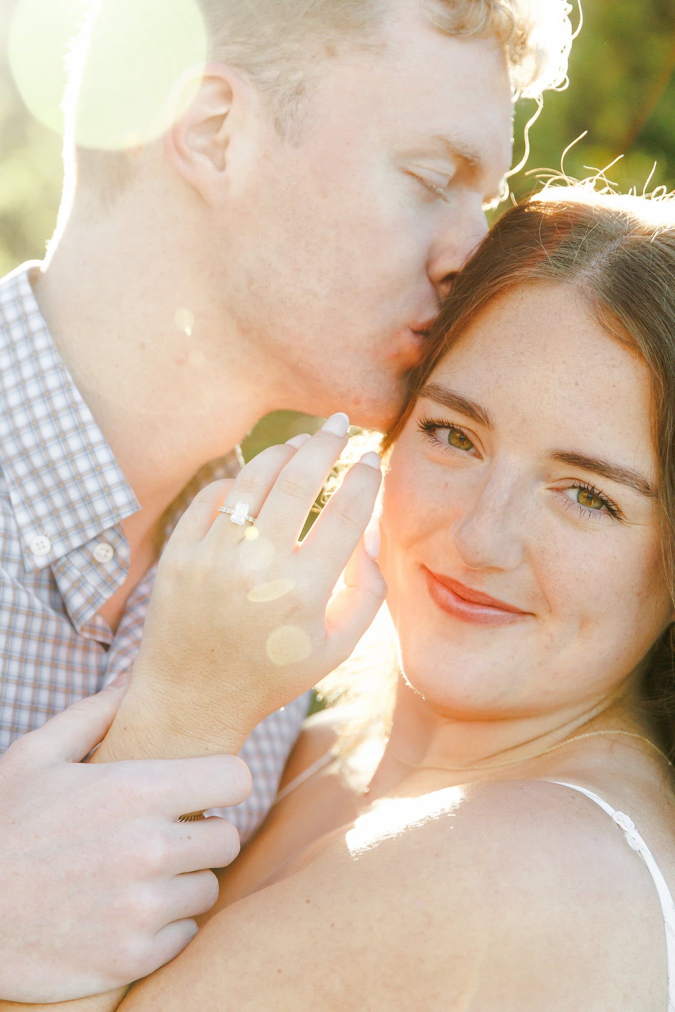 A man kisses a smiling woman's cheek as she shows off her engagement ring in warm golden sunlight.