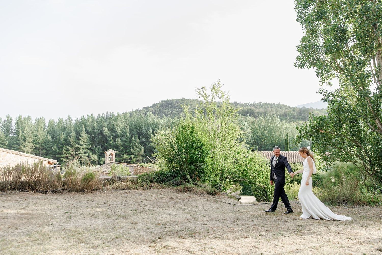 A bride and groom walk hand in hand through a scenic mountain landscape with lush green trees and hills in the background.