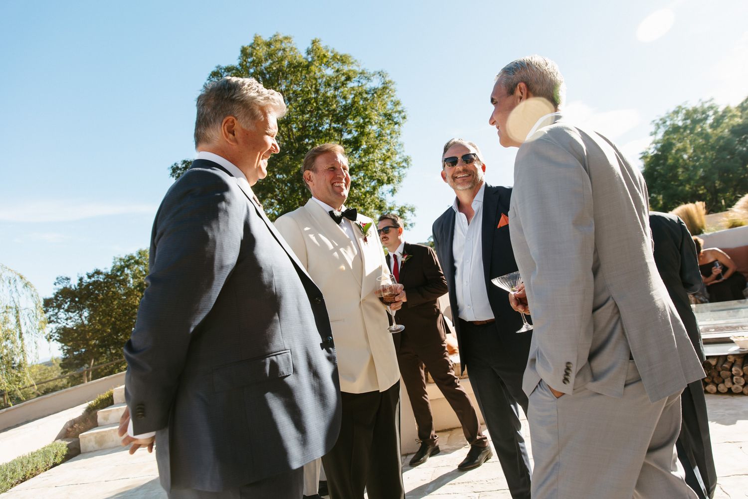 Group of well-dressed men in suits chatting outdoors on a sunny day at an elegant wedding celebration with trees in the background.
